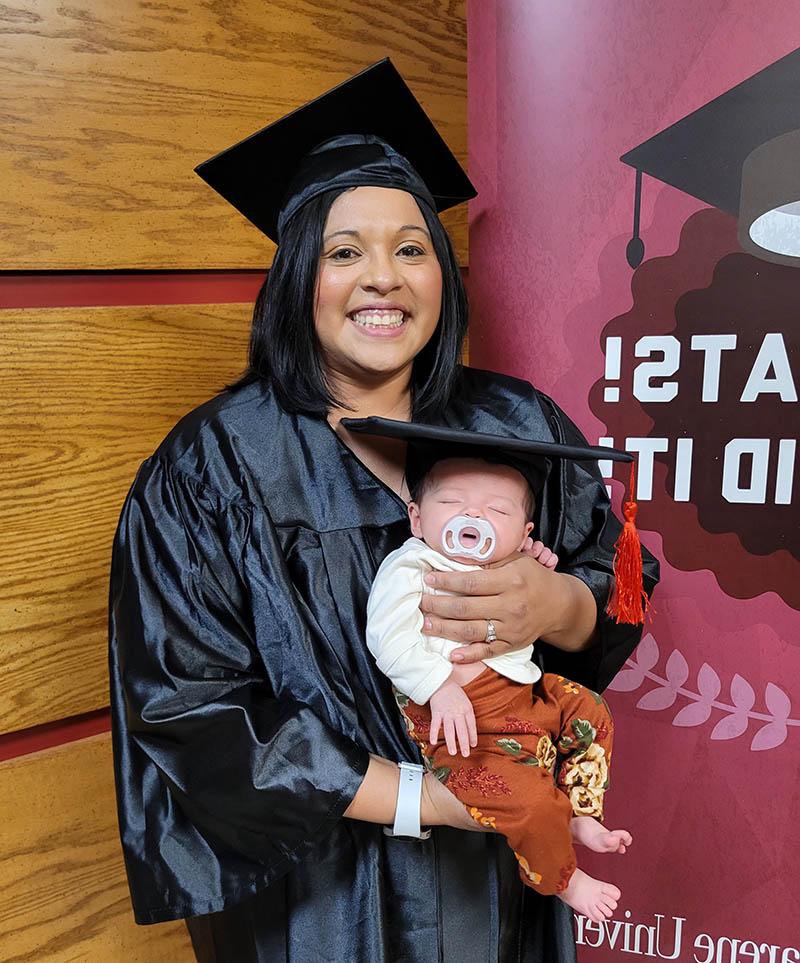 Jessica dressed in her graduation regalia holding her baby